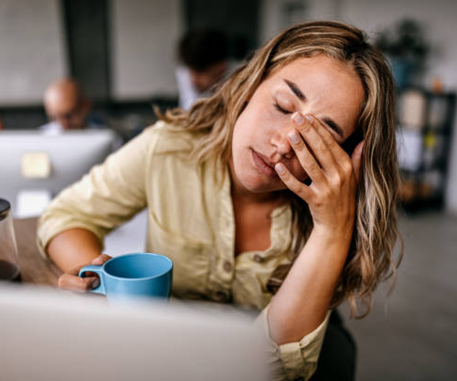 Tired and exhausted young female entrepreneur drinking coffee and rubbing eyes while feeling stressed and worried in coworking space